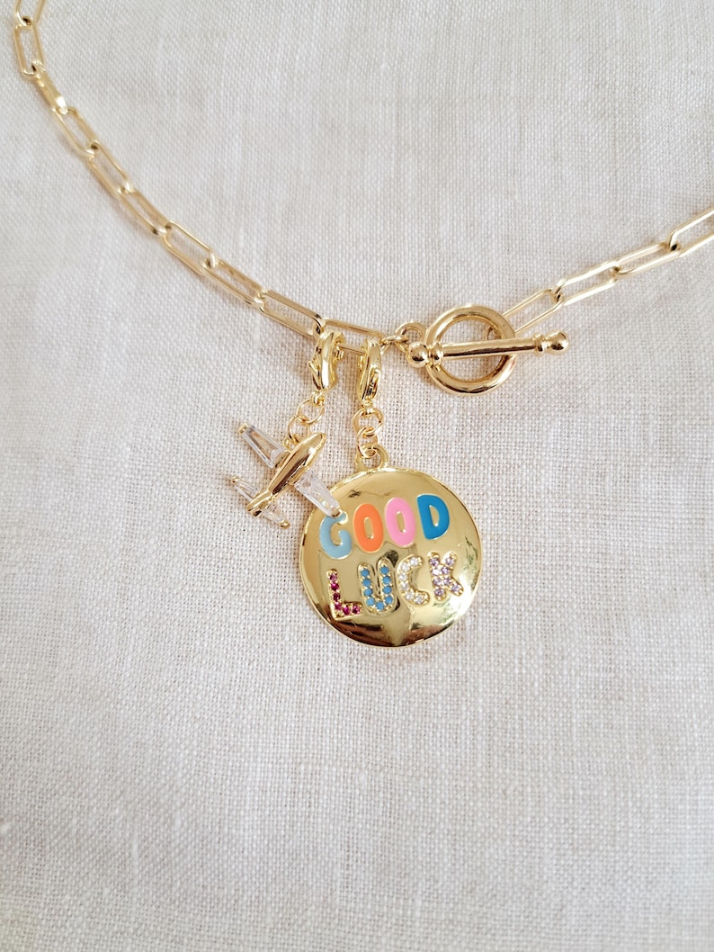 May include: Gold chain necklace with a toggle clasp and two charms. One charm is a small airplane and the other is a round charm with the words "GOOD LUCK" in colorful enamel letters.