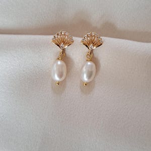 Puede incluir: Un par de pendientes dorados con una parte superior en forma de abanico adornada con pequeñas circonias. Cada pendiente presenta una perla blanca cremosa en forma de lágrima suspendida.