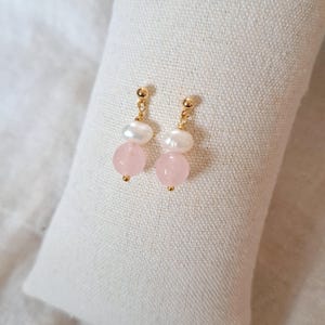 May include: Gold-coloured earrings with a freshwater pearl and a pink rose quartz bead. The earrings are presented on a white, textured surface. The earrings have a post-back closure.