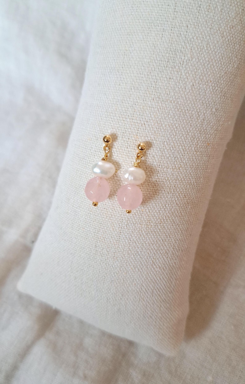 May include: Gold-coloured earrings with a freshwater pearl and a pink rose quartz bead. The earrings are presented on a white, textured surface. The earrings have a post-back closure.