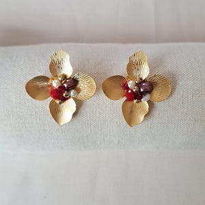 May include: A pair of gold-colored flower-shaped earrings. Each earring features four petals and is adorned with red, white, and purple beads at the center. The earrings are displayed on a white surface.