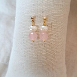 May include: A pair of earrings with gold posts, white pearls, and pink gemstones. The earrings are presented on a white, textured surface. The jewellery has a delicate and feminine aesthetic.