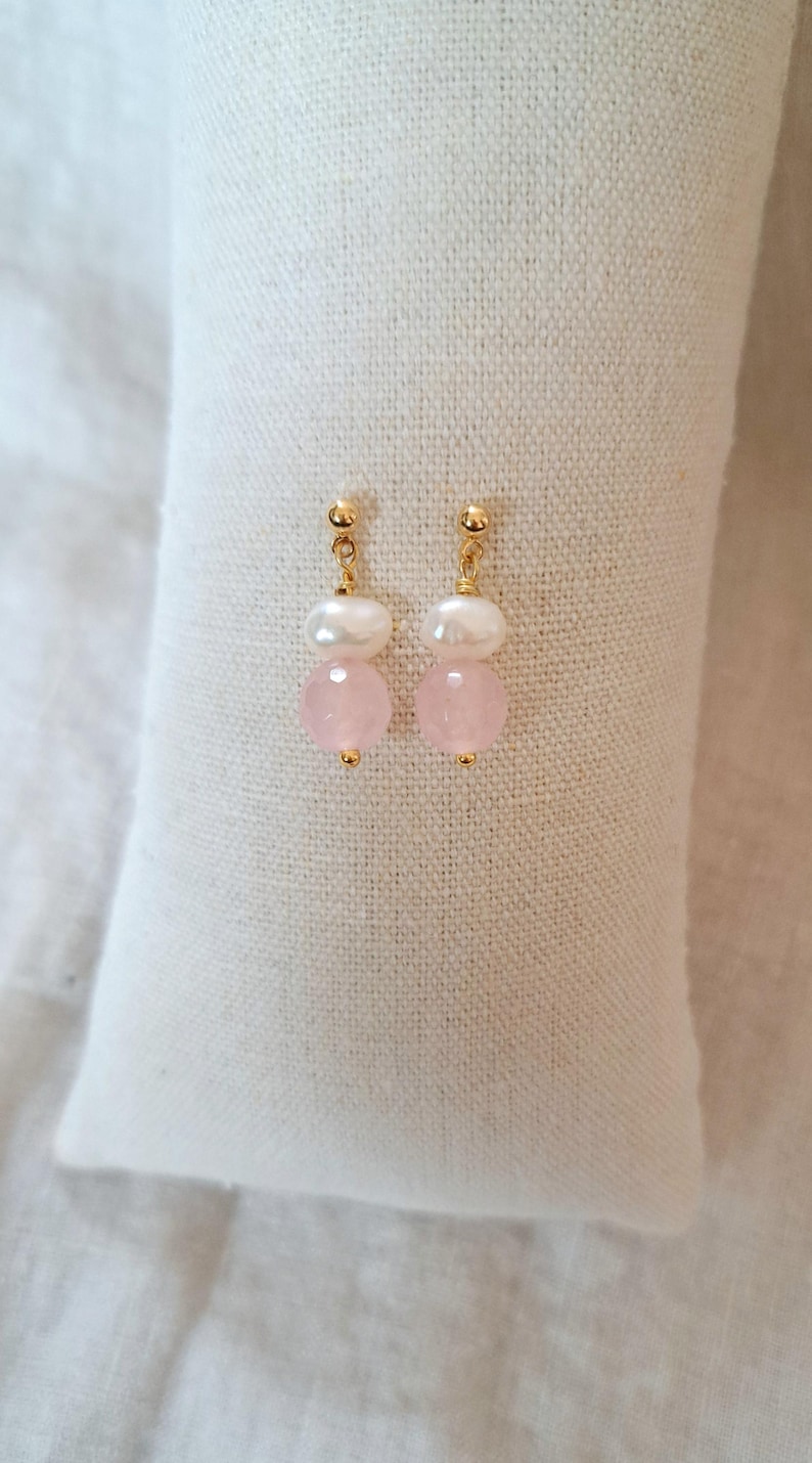 May include: A pair of earrings with gold posts, white pearls, and pink gemstones. The earrings are presented on a white, textured surface. The jewellery has a delicate and feminine aesthetic.