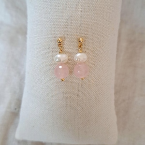 May include: A pair of gold earrings featuring a pearl and a pink gemstone. The earrings are presented on a white, textured surface. The earrings have a post-style fitting.