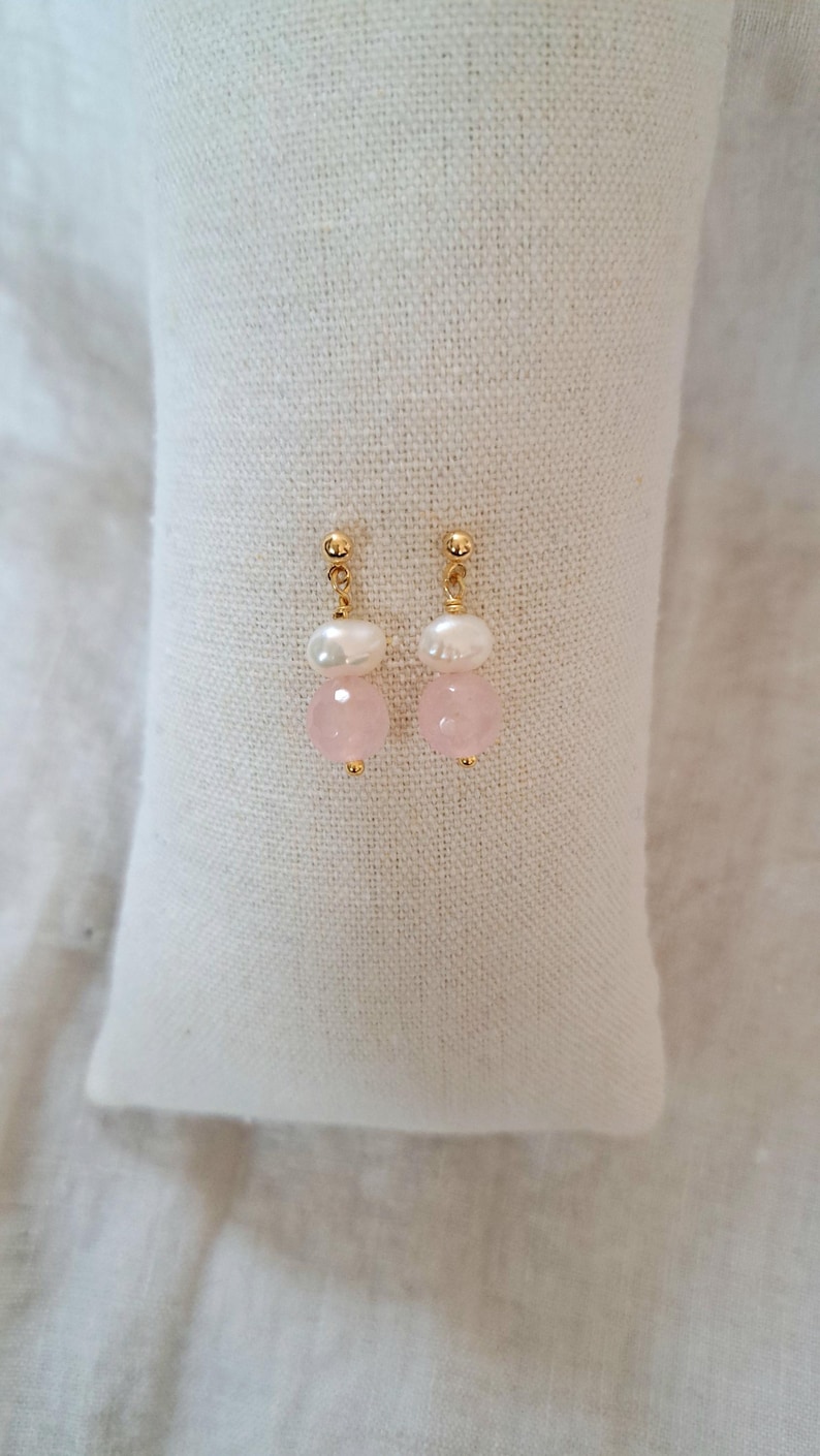May include: A pair of gold earrings featuring a pearl and a pink gemstone. The earrings are presented on a white, textured surface. The earrings have a post-style fitting.