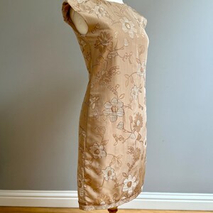 May include: A tan-colored, knee-length dress with a floral pattern. The dress has a round neckline and cap sleeves. The floral design features light and dark brown flowers and leaves. The dress is displayed on a mannequin.