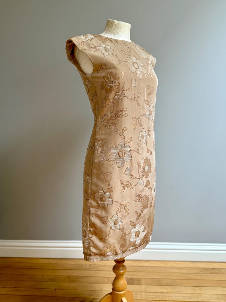 May include: A tan-colored, knee-length dress with a floral pattern. The dress has a round neckline and cap sleeves. The floral design features light and dark brown flowers and leaves. The dress is displayed on a mannequin.