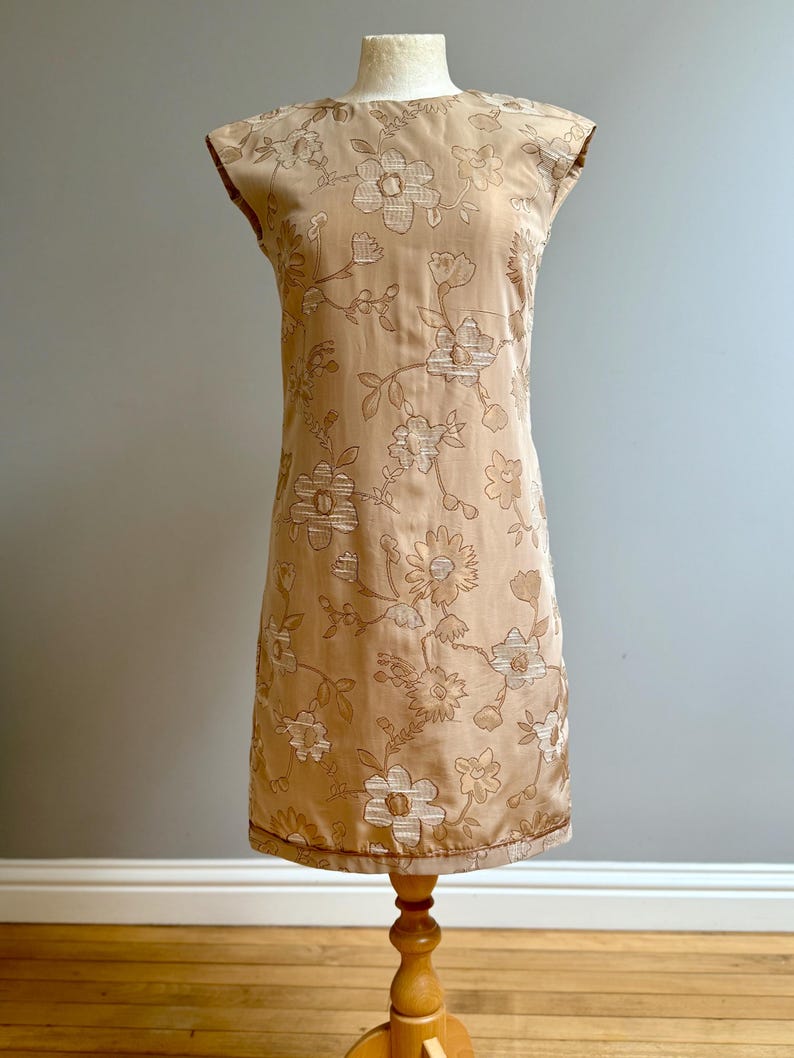 May include: A tan-colored, sleeveless dress with a floral pattern. The dress is displayed on a wooden dress form. The floral design features white and brown embroidered flowers and leaves. The dress has a straight, knee-length cut.