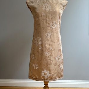 May include: A vintage beige dress with a floral pattern. The sleeveless dress is displayed on a wooden dress form. The dress features a round neckline and a straight silhouette. The floral design is in a lighter beige color.