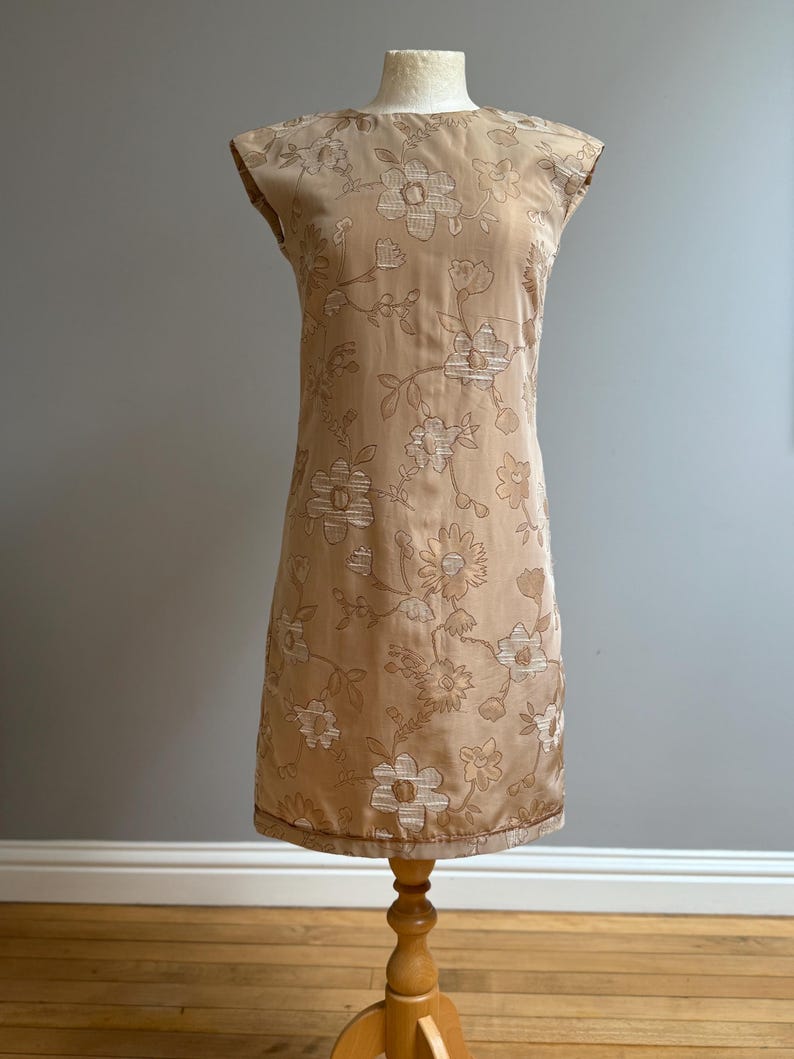 May include: A vintage beige dress with a floral pattern. The sleeveless dress is displayed on a wooden dress form. The dress features a round neckline and a straight silhouette. The floral design is in a lighter beige color.