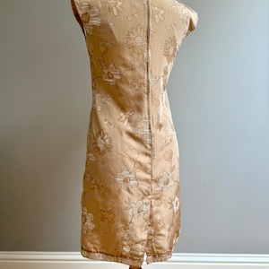 May include: A vintage, knee-length dress in a light tan color, adorned with a floral pattern. The sleeveless dress features a subtle sheen and a slit at the hem. The dress is displayed on a wooden mannequin.