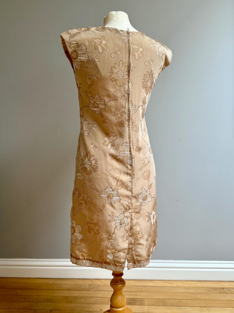 May include: A vintage, knee-length dress in a light tan color, adorned with a floral pattern. The sleeveless dress features a subtle sheen and a slit at the hem. The dress is displayed on a wooden mannequin.