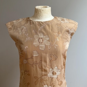 May include: A tan-colored dress with a floral pattern. The dress is sleeveless and has a round neckline. The floral pattern is in a lighter tan color and covers the entire dress. The dress is displayed on a mannequin.