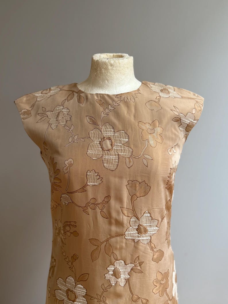 May include: A tan-colored dress with a floral pattern. The dress is sleeveless and has a round neckline. The floral pattern is in a lighter tan color and covers the entire dress. The dress is displayed on a mannequin.