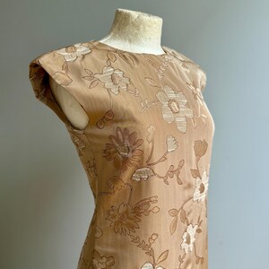 May include: A tan-colored dress with a floral pattern. The dress features a round neckline and cap sleeves. The floral design is embroidered in a lighter tan color, creating a subtle contrast. The dress is displayed on a mannequin.