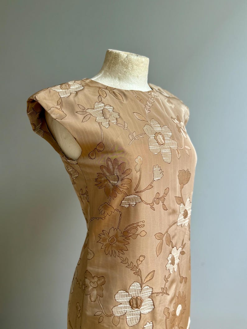 May include: A tan-colored dress with a floral pattern. The dress features a round neckline and cap sleeves. The floral design is embroidered in a lighter tan color, creating a subtle contrast. The dress is displayed on a mannequin.
