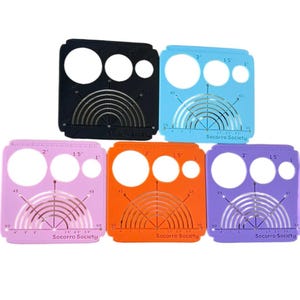 Könnte beinhalten: Fünf kreisförmige Kunststoffschablonen in Schwarz, Blau, Rosa, Orange und Lila. Jede Schablone hat drei kreisförmige Aussparungen, Gradmarkierungen und den Text "Socorro Society". Die Schablonen messen 5,08 cm, 3,81 cm und 2,54 cm.