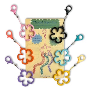 May include: A collection of colorful flower-shaped thread organizers with attached clips. The organizers come in pink, orange, purple, black, blue, yellow, and teal. A cardboard display with the text "Thread Organizer" is in the background.