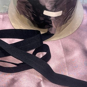 May include: A roll of black elastic ribbon sits on a cardboard base, with a length of the ribbon unrolled and lying on a pink satin surface. The ribbon is a solid black color and appears to be made of a soft, flexible material.