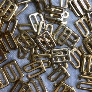 Puede incluir: Un montón de reguladores de tirantes de sujetador de metal dorado. Los reguladores son de forma rectangular con dos agujeros a cada lado.