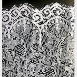 May include: White lace fabric with a delicate floral pattern. The lace has a scalloped edge and a repeating design of small circles and swirls.