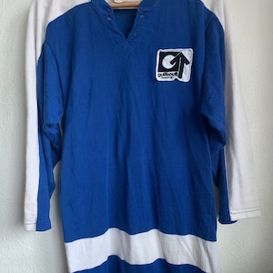 Op de afbeelding: Een blauw en wit langarm shirt met een witte streep aan de onderkant en een patch op de borst met de tekst "Outback Transport" met een pijl die omhoog wijst.