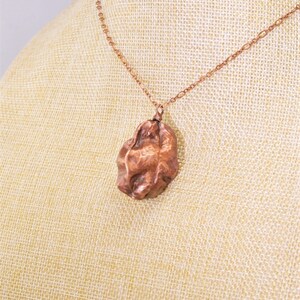 Könnte beinhalten: Eine Kupferkette mit einem strukturierten, organisch geformten Anhänger. Der Anhänger ist dunkelkupferfarben und hat eine raue, gehämmerte Oberfläche.