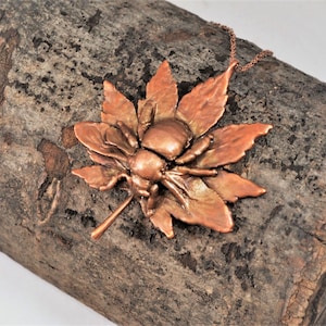 May include: A copper necklace featuring a stylized bee on a maple leaf. The necklace is displayed on a piece of weathered wood.