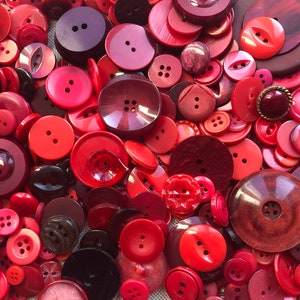 May include: A collection of various red buttons in different sizes and shades, including crimson, scarlet, and burgundy. The buttons have two or four holes, and some have a shank. The assortment includes round, square, and decorative shapes, creating a textured, colorful display.