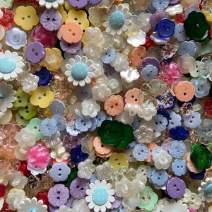 Puede incluir: Primer plano de una colección de botones decorativos en varios colores y formas. Los botones incluyen diseños de flores, nubes y redondos en tonos de rosa, azul, amarillo, verde y morado. Algunos tienen un acabado nacarado.