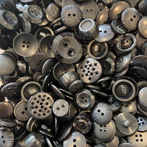 May include: A close-up of a pile of black buttons in various shapes and sizes. Some buttons have two or four holes, others have textured surfaces, star patterns, or ridged edges. The buttons are made of a shiny, dark material.