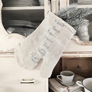 Può includere: Un guanto da forno grigio chiaro con la scritta "Gritti" stampata in grigio scuro. Il guanto è appeso a un gancio di metallo su un mobile bianco. Un rametto di sempreverde è infilato all'interno. Stoviglie visibili sullo sfondo.
