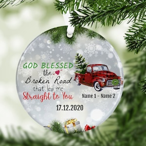May include: A round, white Christmas ornament with a red truck carrying a Christmas tree. The ornament has a snowy background with snowflakes and the text "GOD BLESSED the Broken Road that led me Straight to You Name 1 - Name 2 17.12.2020".