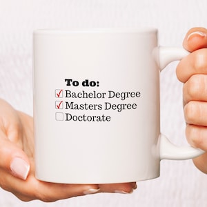 May include: White ceramic coffee mug with black text that reads "To do:" and a checklist with the following items: "Bachelor Degree", "Masters Degree", and "Doctorate". The first two items are checked off with red tick marks.