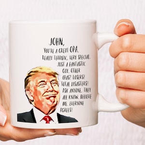 May include: White ceramic mug with a black and white illustration of Donald Trump. The mug has the text "JOHN, YOU'RE A GREAT OPA, REALLY TERRIFIC, VERY SPECIAL. JUST A FANTASTIC GUY. OTHER OPAS? LOSERS! TOTAL DISASTERS! ASK ANYONE, THEY ALL KNOW. BELIEVE ME. EVERYONE AGREES!"