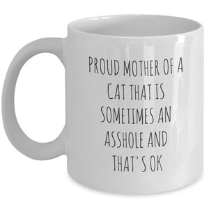 May include: White ceramic mug with a curved handle. The mug features black text that reads: "PROUD MOTHER OF A CAT THAT IS SOMETIMES AN ASSHOLE AND THAT'S OK."