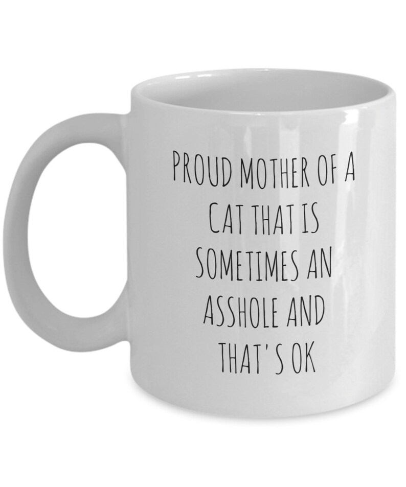 May include: White ceramic mug with a curved handle. The mug features black text that reads: "PROUD MOTHER OF A CAT THAT IS SOMETIMES AN ASSHOLE AND THAT'S OK."