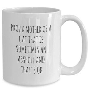 May include: White ceramic mug with a black text that reads "PROUD MOTHER OF A CAT THAT IS SOMETIMES AN ASSHOLE AND THAT'S OK". The mug has a curved handle.