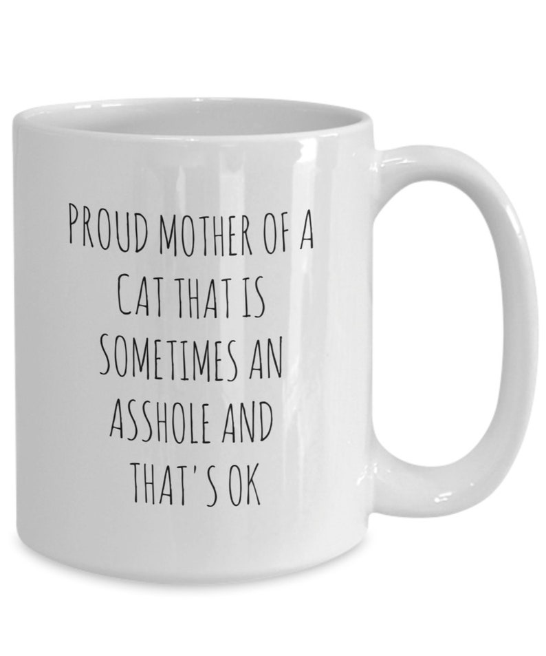 May include: White ceramic mug with a black text that reads "PROUD MOTHER OF A CAT THAT IS SOMETIMES AN ASSHOLE AND THAT'S OK". The mug has a curved handle.