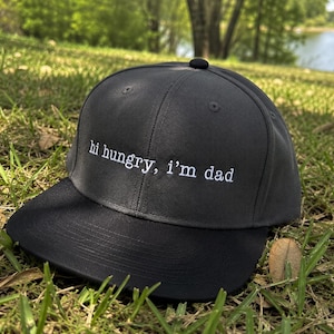 K&ouml;nnte beinhalten: Eine schwarze Baseballkappe mit gebogenem Schirm, auf gr&uuml;nem Gras. Auf der Kappe steht in wei&szlig;er Schrift "hi hungry, i'm dad". Ein l&auml;ssiges Accessoire f&uuml;r den Alltag.