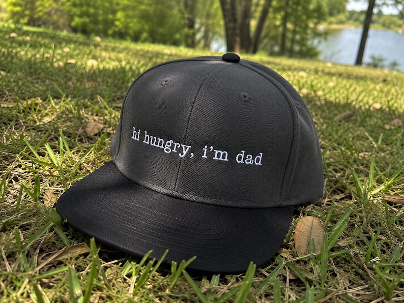 K&ouml;nnte beinhalten: Eine schwarze Baseballkappe mit gebogenem Schirm, auf gr&uuml;nem Gras. Auf der Kappe steht in wei&szlig;er Schrift "hi hungry, i'm dad". Ein l&auml;ssiges Accessoire f&uuml;r den Alltag.