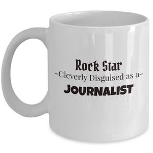 May include: White ceramic mug with a curved handle. The mug features the text "Rock Star - Cleverly Disguised as a- JOURNALIST" in black, bold font. The mug is a standard size for coffee or tea.