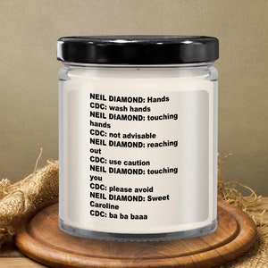 May include: A white candle jar with a black lid and a white label with black text. The text on the label reads: "NEIL DIAMOND: Hands CDC: wash hands NEIL DIAMOND: touching hands CDC: not advisable NEIL DIAMOND: reaching out CDC: use caution NEIL DIAMOND: touching you CDC: please avoid NEIL DIAMOND: Sweet Caroline CDC: ba ba baaa"