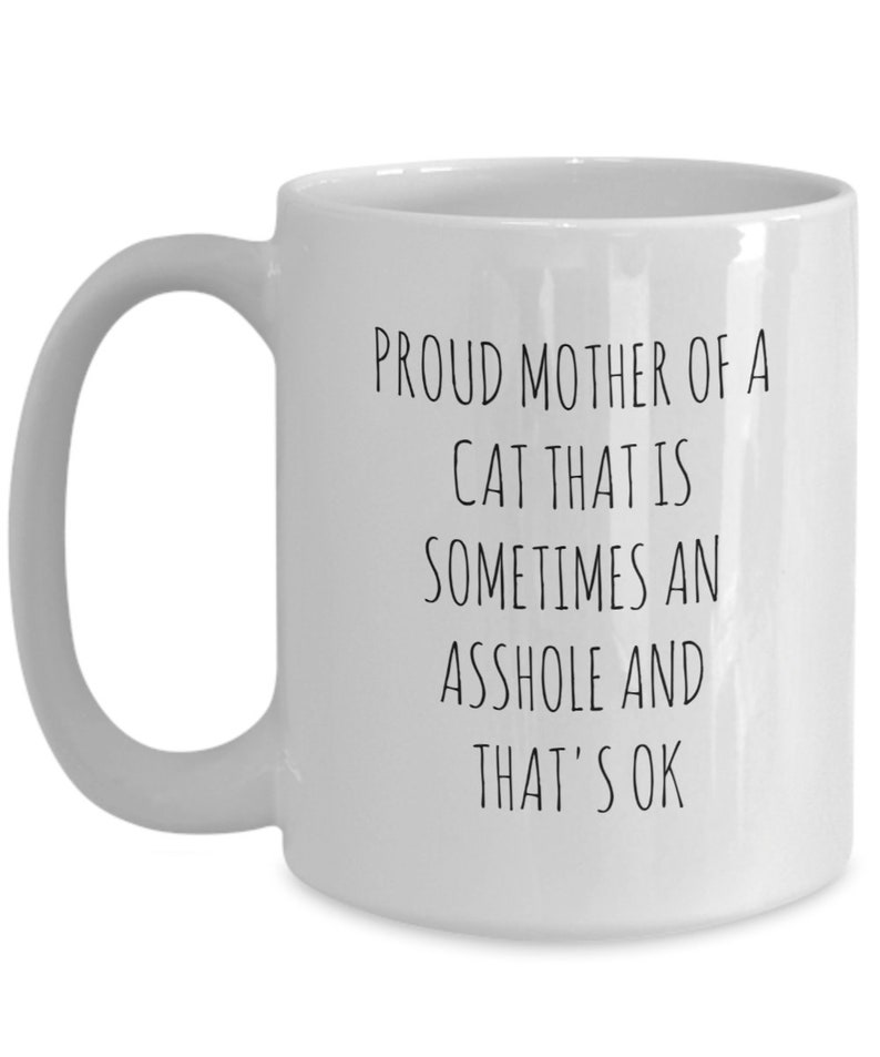 May include: White ceramic mug with a curved handle. The mug features black text that reads: "PROUD MOTHER OF A CAT THAT IS SOMETIMES AN ASSHOLE AND THAT'S OK."