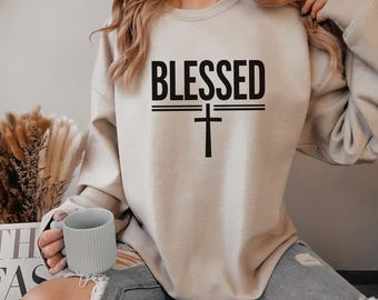 May include: A beige sweatshirt with the word "BLESSED" printed in black letters and a black cross underneath. The sweatshirt is being worn by a person.
