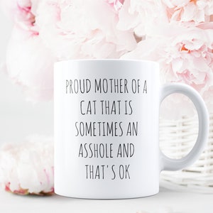 May include: White ceramic mug with the text "PROUD MOTHER OF A CAT THAT IS SOMETIMES AN ASSHOLE AND THAT'S OK" in black lettering. The mug is set against a backdrop of pink flowers and a white wicker basket.