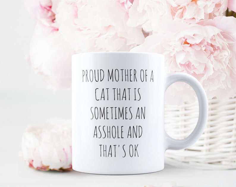 May include: White ceramic mug with the text "PROUD MOTHER OF A CAT THAT IS SOMETIMES AN ASSHOLE AND THAT'S OK" in black lettering. The mug is set against a backdrop of pink flowers and a white wicker basket.
