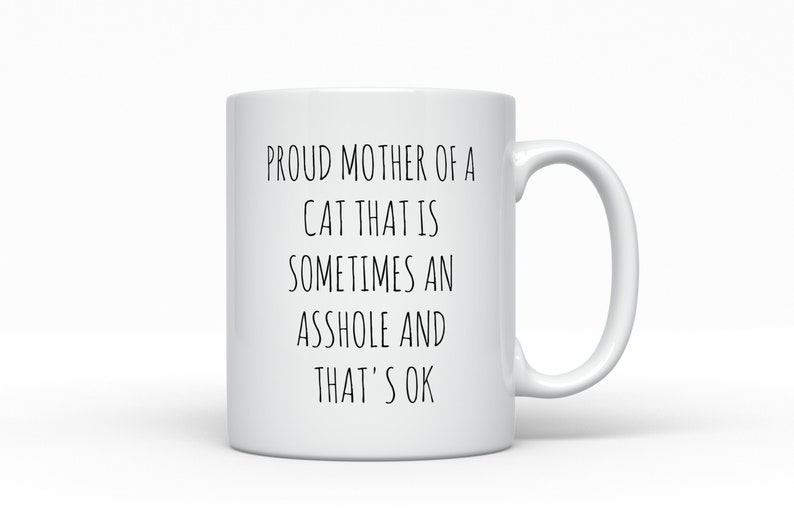 May include: White ceramic mug with a black handle. The mug features the text: "PROUD MOTHER OF A CAT THAT IS SOMETIMES AN ASSHOLE AND THAT'S OK" in black, centered on the mug. The mug is set against a white background.