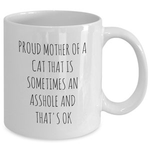 May include: White ceramic mug with a curved handle. The mug features black text that reads: "PROUD MOTHER OF A CAT THAT IS SOMETIMES AN ASSHOLE AND THAT'S OK."