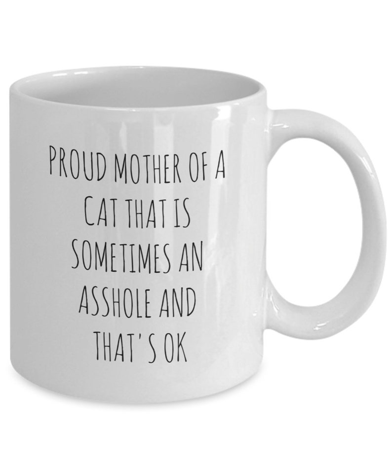 May include: White ceramic mug with a curved handle. The mug features black text that reads: "PROUD MOTHER OF A CAT THAT IS SOMETIMES AN ASSHOLE AND THAT'S OK."